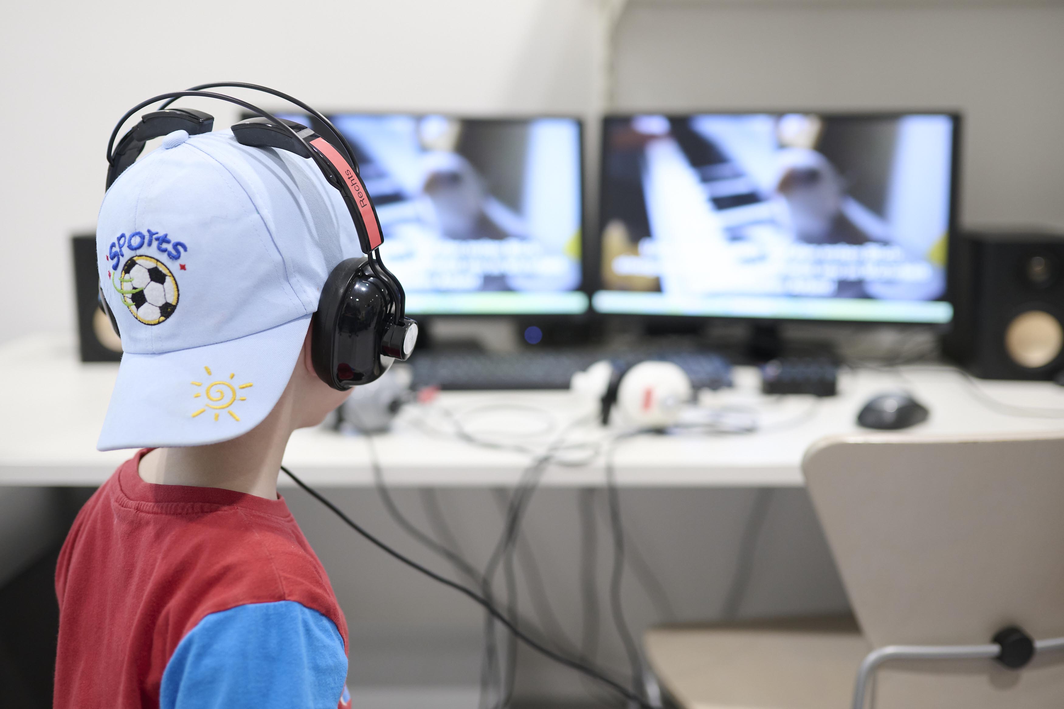 A child wearing a baseball cap and headphones sits at a computer, seen from behind.