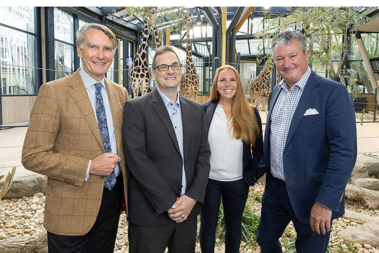 Angela Stöger-Horwath standing with the Rector and a Dean of the University of Vienna, and the Director of Tiergarten Schönbrunn, inside a giraffe enclosure with giraffes visible in the background.