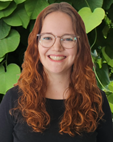 Portrait of Melina Witt with long, curly red hair and glasses, smiling in front of a leafy green background