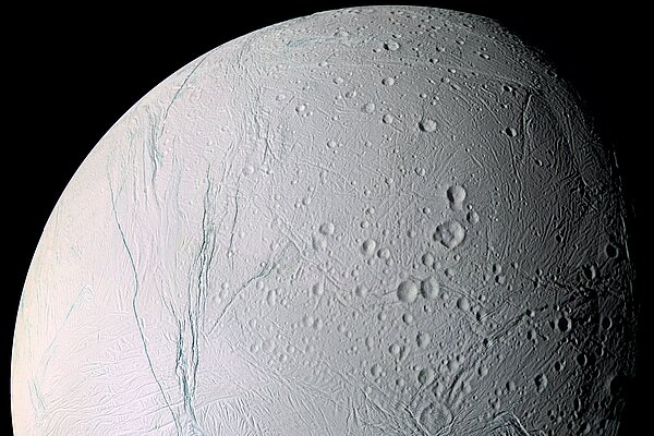 Nahaufnahme des eisigen Saturnmondes Enceladus, fotografiert von der Raumsonde Cassini. Zu sehen ist eine helle, von Kratern und tiefen Furchen durchzogene Eisoberfläche vor dem schwarzen Weltraum.