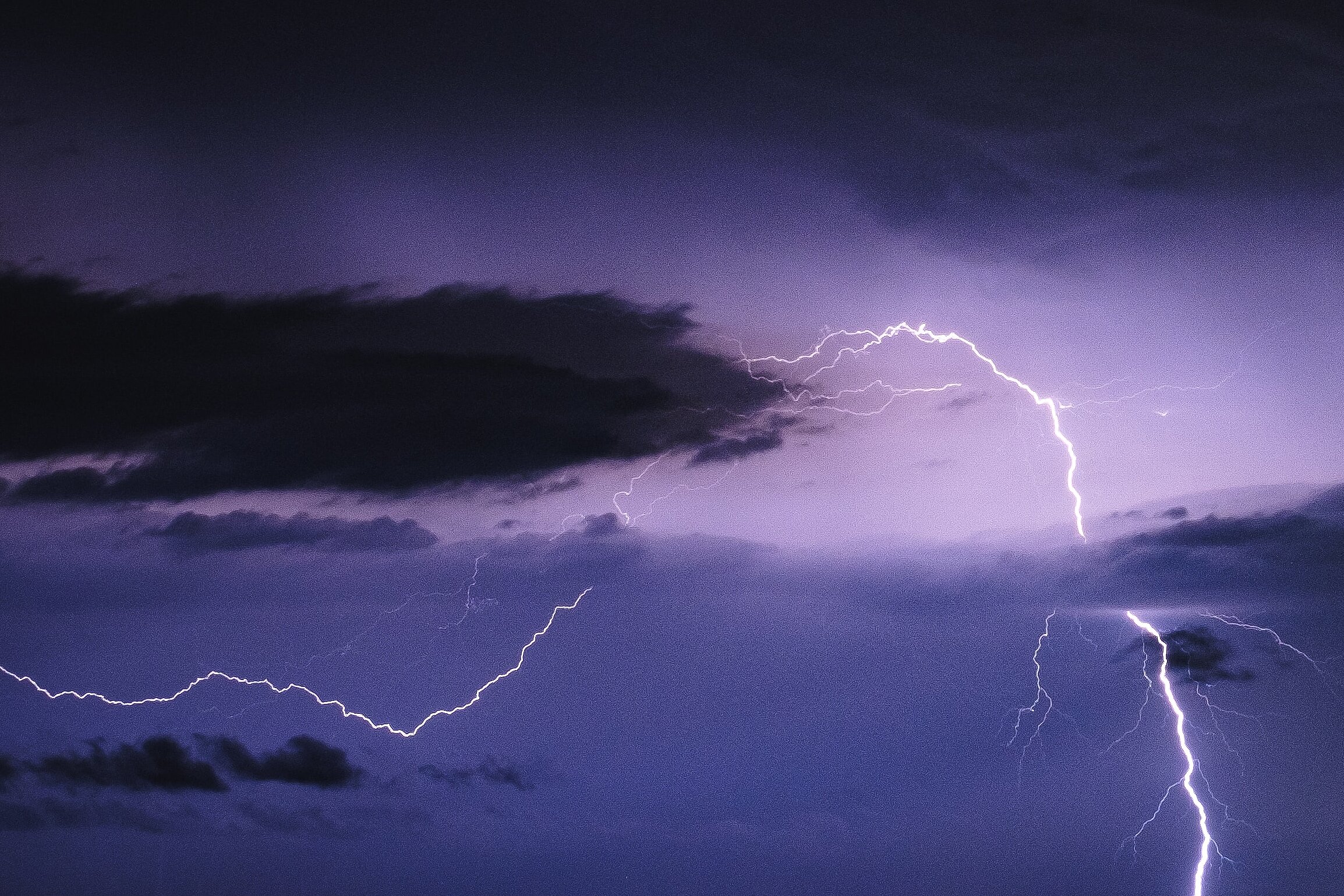 Ein heller Blitz zuckt durch einen dunkelvioletten Nachthimmel voller Gewitterwolken.