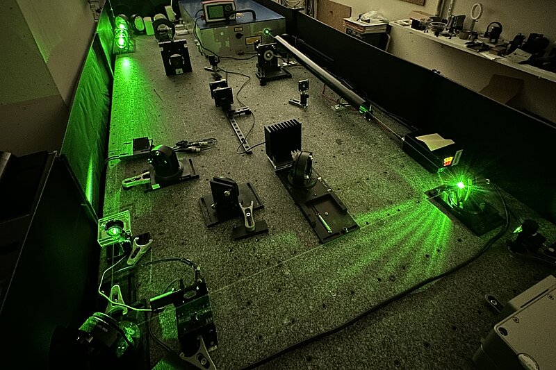 Blick von oben auf einen optischen Labortisch mit verschiedenen Halterungen, Spiegeln und Linsen. Ein intensiv leuchtender, grüner Laserstrahl wird in mehrere Pfade aufgeteilt und reflektiert.Blick von oben auf einen optischen Labortisch mit verschiedenen Halterungen, Spiegeln und Linsen. Ein intensiv leuchtender, grüner Laserstrahl wird in mehrere Pfade aufgeteilt und reflektiert.