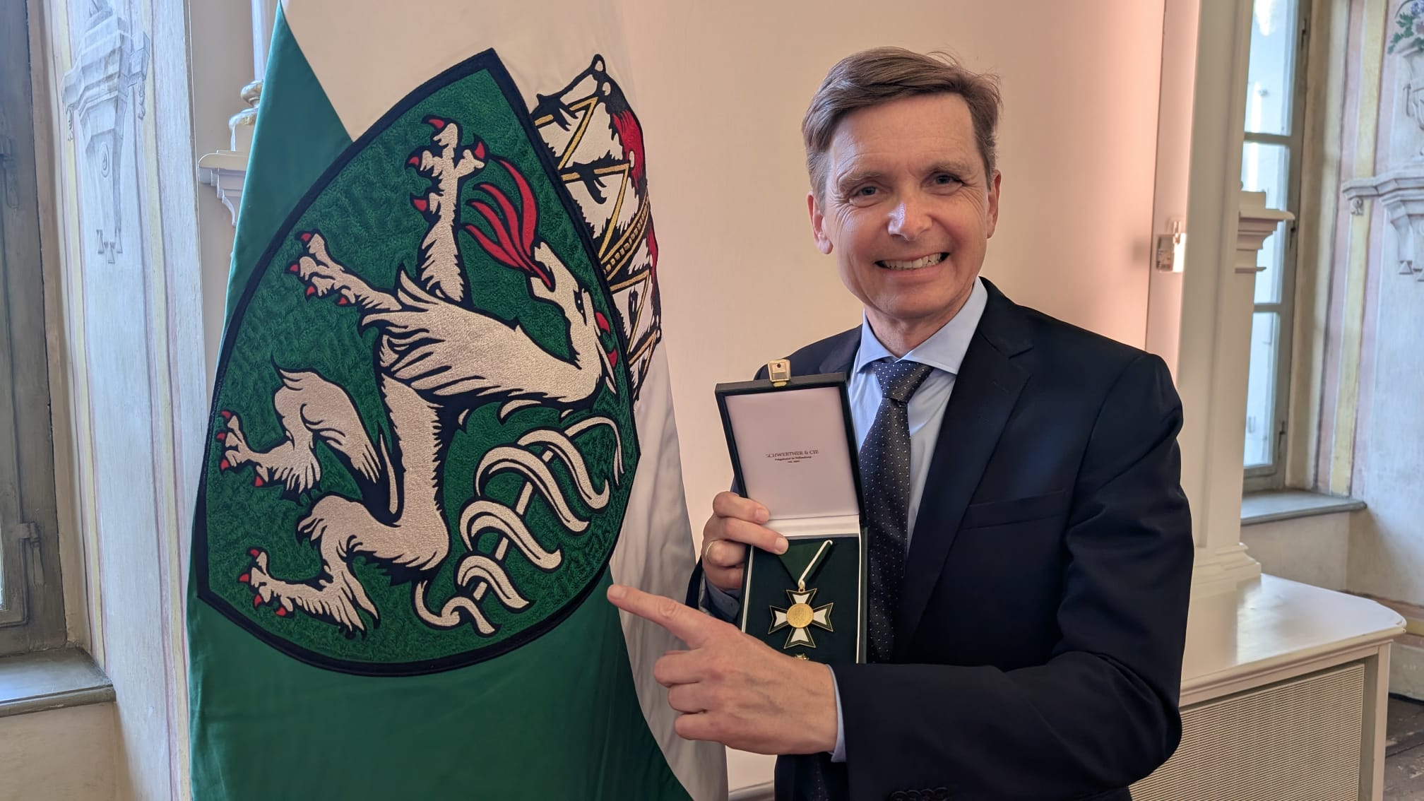Werner Magnes zeigt mit der linken Hand auf die steirische Flagge und hält das Ehrenzeichen des Landes Steiermark für Wissenschaft, Forschung und Kunst in seiner rechteh Hand.