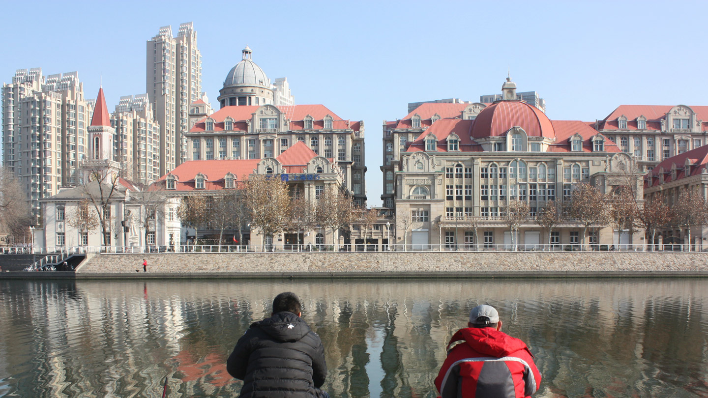Zwei Menschen sitzen an einem städtischen Kanal, hinter dem sich historische Prachtbauten erheben