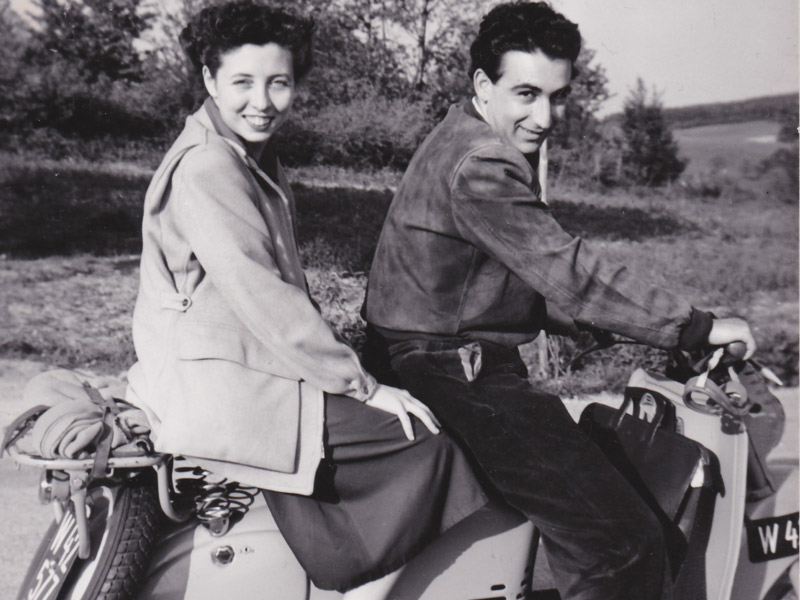 Eine Frau und ein Mann sitzen vor einem ländlichen Hintergrund auf einem Moped