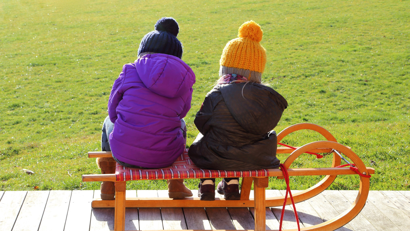 Zwei Kinder in Schianzügen sitzen vor einer grünen Wiese auf einer Rodel