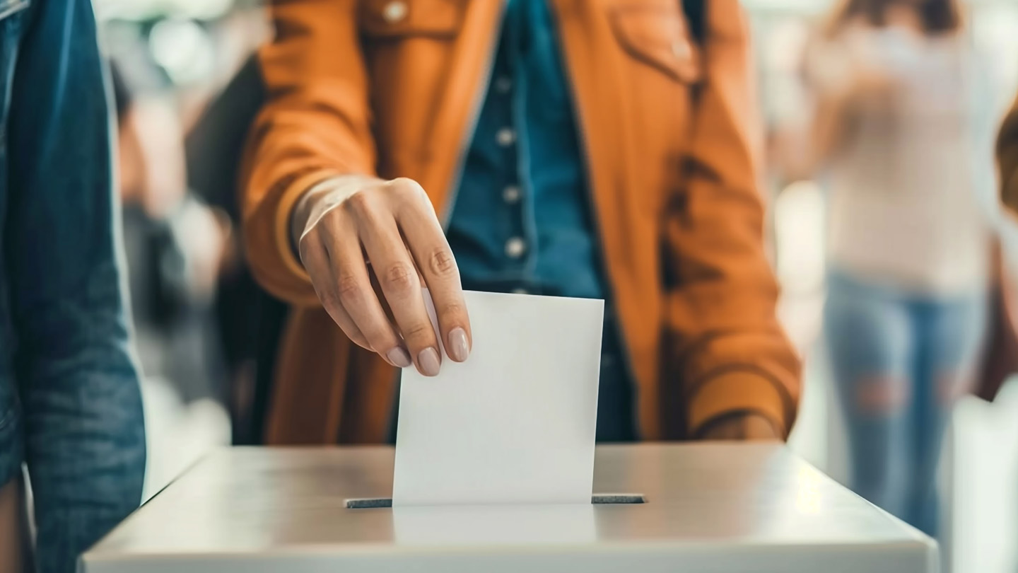 Ein Mensch, dessen Kopf nicht im Bild ist, steckt einen Wahlschein in eine Wahlurne