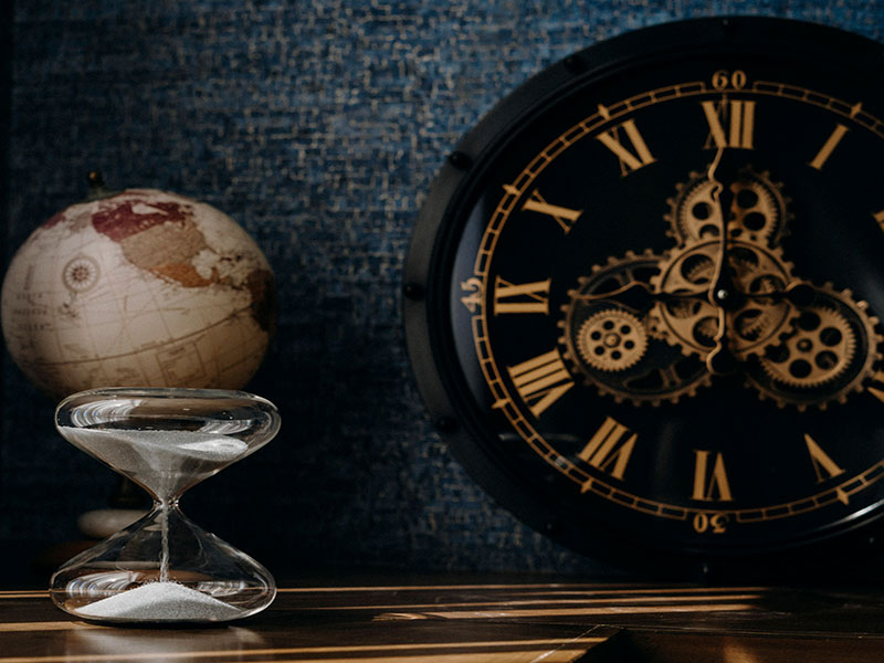 Eine Sanduhr mit fließendem Sand steht auf einem Holztisch. Im Hintergrund sind eine Weltkugel und eine große, schwarze Wanduhr mit römischen Ziffern und sichtbaren Zahnrädern zu sehen.
