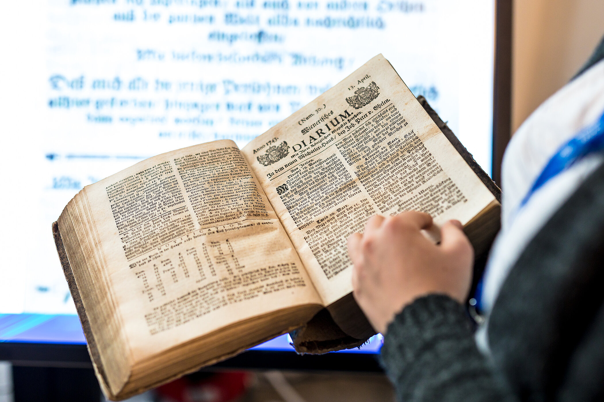 Someone is holding a bound version of the Diarium, opened to a title page, in front of a large screen with a digitized version of a different page.
