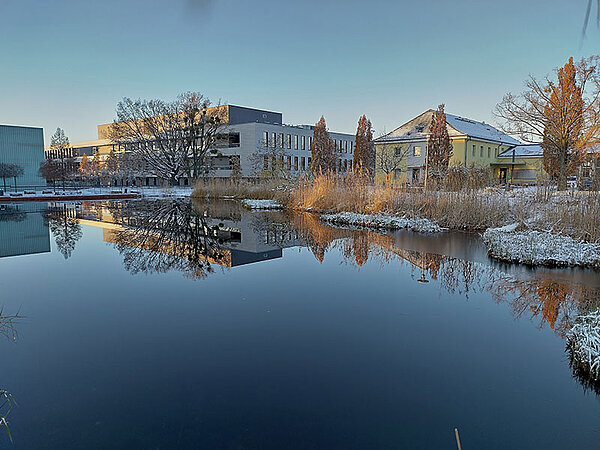 Ansicht eines modernen Gebäudekomplexes und eines historischen Gebäudes, die sich in einem ruhigen Teich spiegeln. Umgeben von kahlen Bäumen und Schilf, deutet die frostige Landschaft auf eine winterliche Atmosphäre hin.