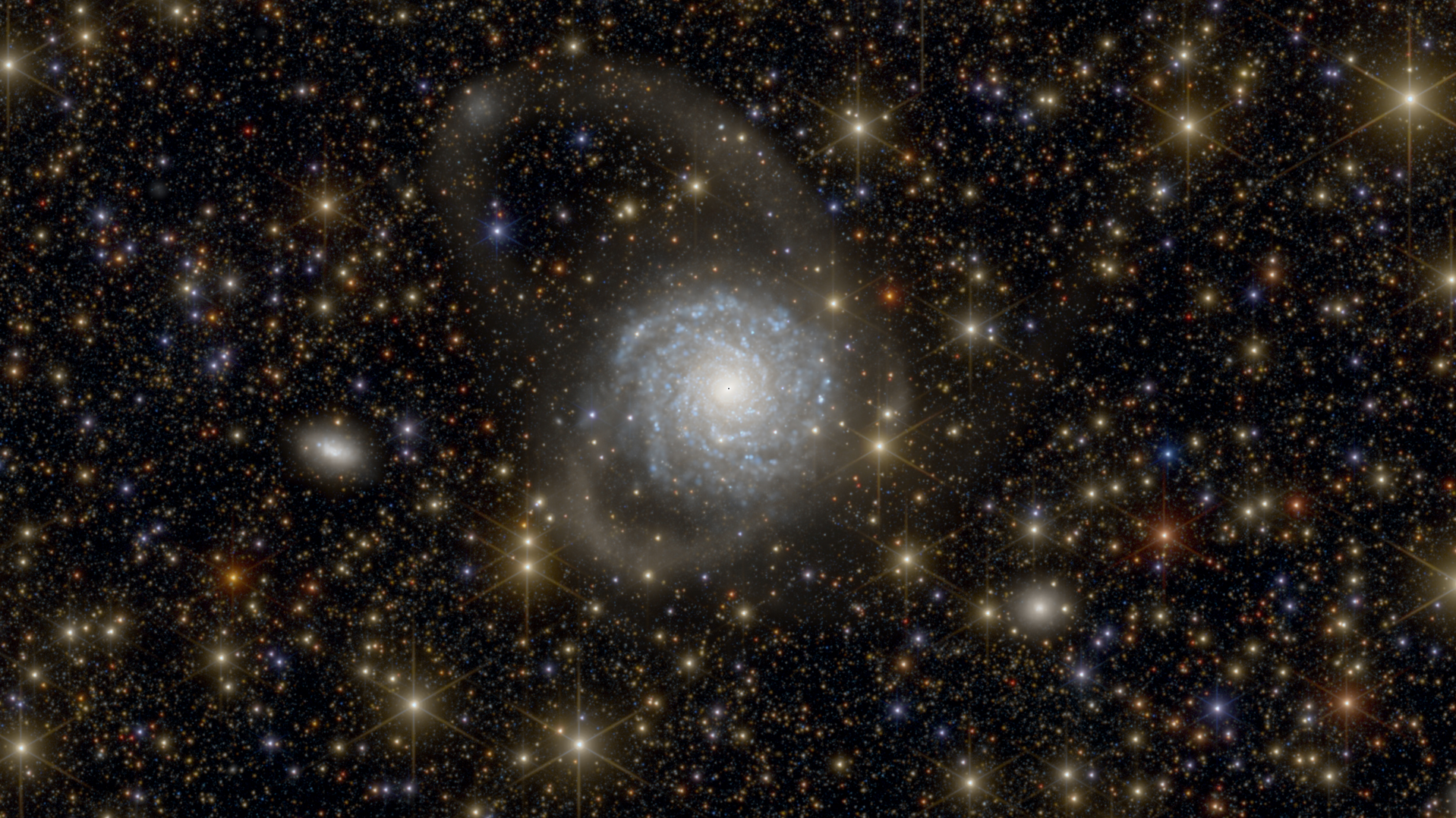 A blue-white, circular spiral galaxy is shown on a colourful starry background. Two faint, cloud-like arcs extend from the galaxy's central disc: one U-shaped loop sticks out to the lower left, and a larger hook-shaped arc extends up and to the left. A diffusely glowing oval-shaped dwarf galaxy lies to the left of the larger galaxy.