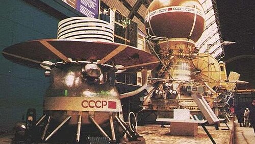 Two historic metal space probes bearing the inscription “CCCP” are on display in a large hall. The probe in the foreground has a wide, circular shield on its top, while the model behind it features a large sphere.