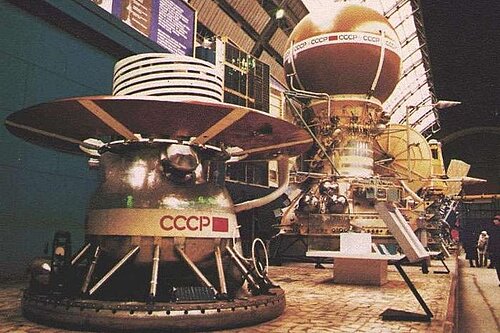 Two historic metal space probes bearing the inscription “CCCP” are on display in a large hall. The probe in the foreground has a wide, circular shield on its top, while the model behind it features a large sphere.