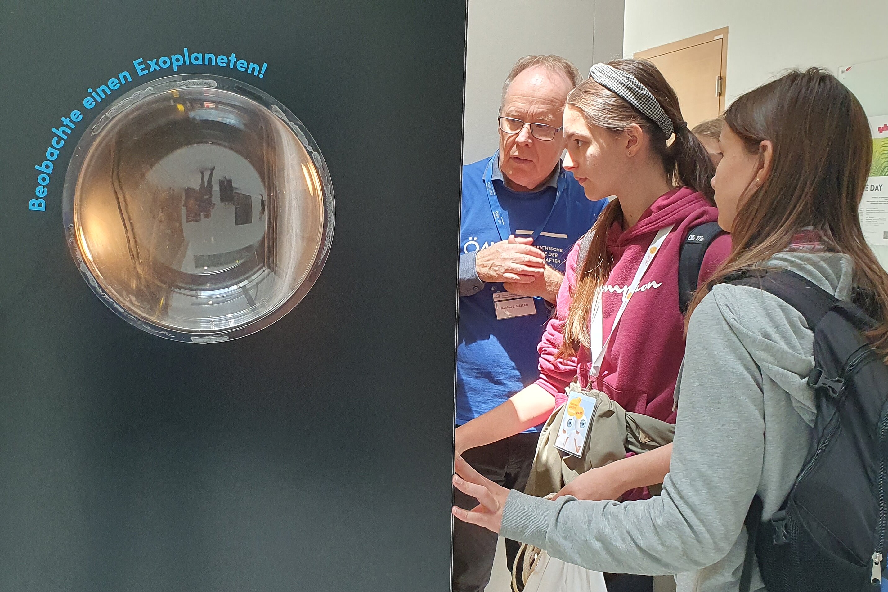 On the left side, there is a big black box with a round window and the text "Observe an exoplanet". On the other side there are standing a few teenagers and an adult who explains the exhibit to them.