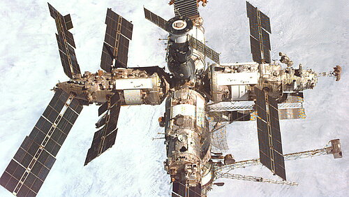 A large, cross-shaped space station with numerous modules and dark solar panel wings is shown from directly above. In the background, the Earth's surface stretches out beneath a bright, nearly continuous cloud cover.