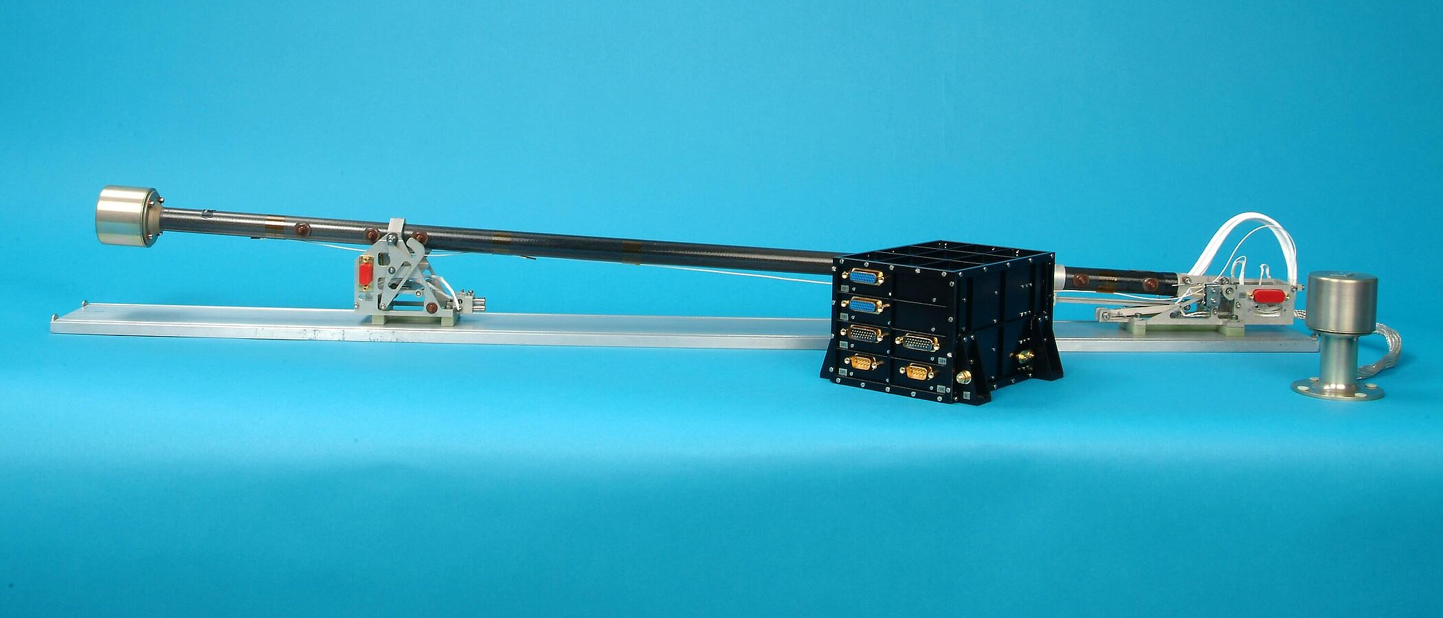 A technical assembly consisting of a long, thin black rod is mounted horizontally on a metal rail. In front of the rod is a small black box with several gold-colored connectors against a light blue background.