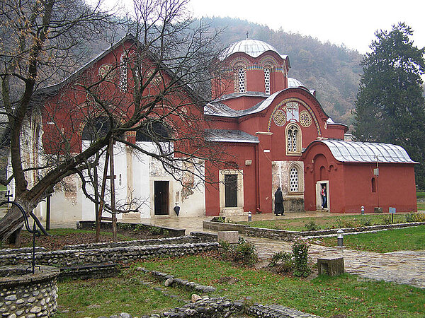Rötlich verputzte orthodoxe Klosterkirche Patriarchat Peć mit weißem Anbau, Kuppel und dekorativen Fensterfassungen; davor stehen kahle Bäume und ein niedriger Steinmauerrest. Im Hintergrund erhebt sich ein bewaldeter Hügel.
