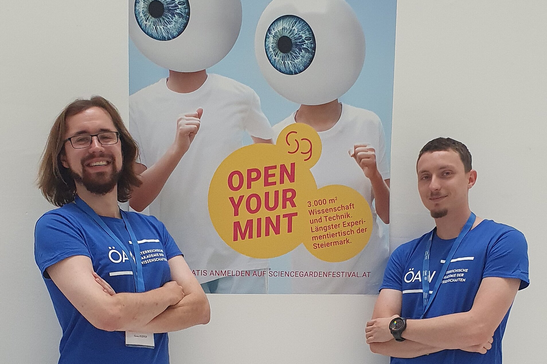 On the image, you can see two men standing in front of a "science garden festival" poster, which is hung on a white wall. Both men wear blue T-Shirts that say "ÖAW".