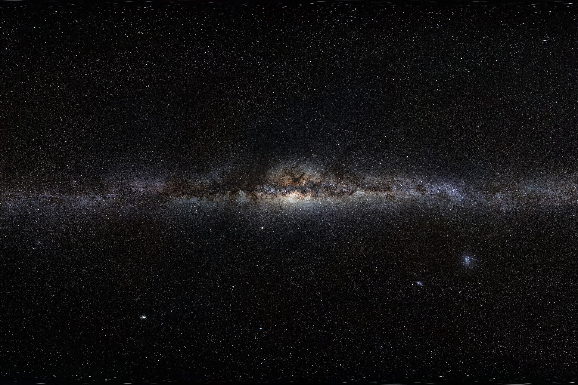 Das Bild zeigt die Milchstraße als langes, helles Band mitten im dunklen Sternenhimmel. In der Mitte leuchtet die Galaxie besonders stark, und weiter unten sind zwei kleine Nachbargalaxien zu sehen.