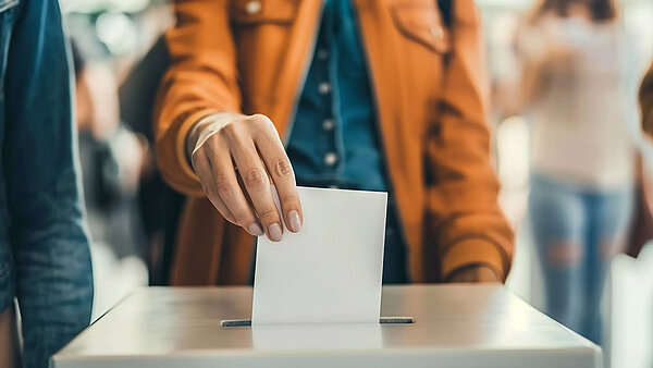 Ein Mensch, dessen Kopf nicht im Bild ist, steckt einen Wahlschein in eine Wahlurne