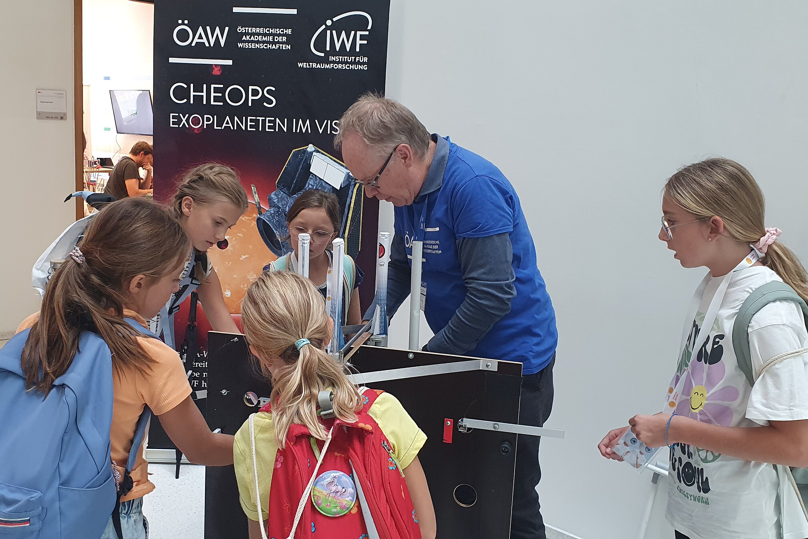 A group of girls is standing around a display at an exhibition where a miniature rocket is being launched. There is also a banner that reads “CHEOPS: Targeting Exoplanets.”