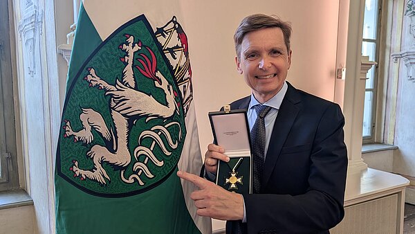 Werner Magnes zeigt mit der linken Hand auf die steirische Flagge und hält das Ehrenzeichen des Landes Steiermark für Wissenschaft, Forschung und Kunst in seiner rechteh Hand.