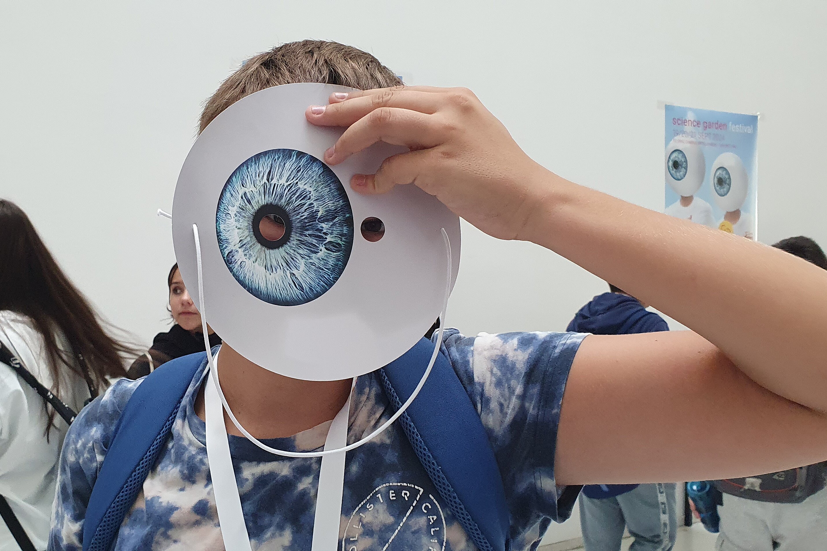 The picture shows a boy holding a mask in front of his face, which looks like a huge eye with a blue iris.