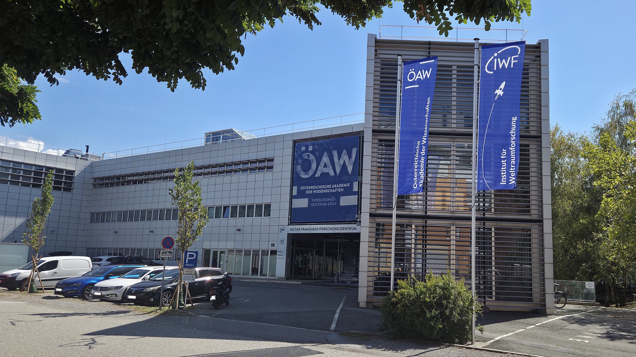 An exterior view of a modern research building with a striking glass facade on the right and a large blue banner reading “ÖAW” above the entrance. In front of the building is a paved forecourt with a few small trees under a slightly cloudy sky.