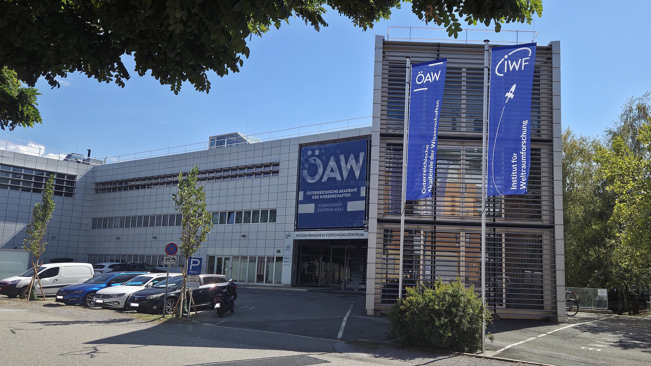 An exterior view of a modern research building with a striking glass facade on the right and a large blue banner reading “ÖAW” above the entrance. In front of the building is a paved forecourt with a few small trees under a slightly cloudy sky.