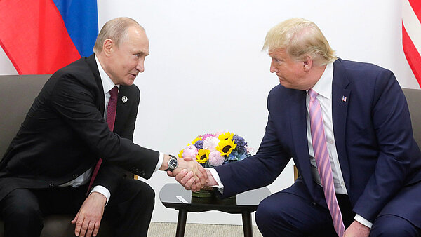 Wladimir Putin und Donald Trump sitzen nebeneinander und reichen sich vor einem Blumenstrauß die Hand.