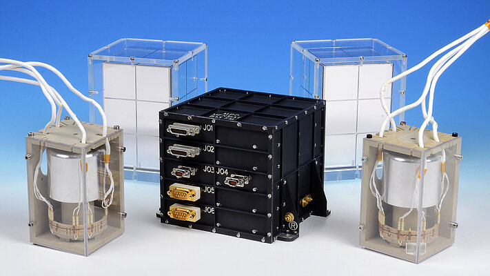 The image shows several parts of a space instrument against a blue background. In the center is a black box, with two identical silver sensors on either side of it. Thick white cables extend upward from the sensors.
