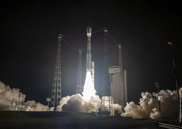 A night-time photo showing a rocket taking off from a launchpad. It is surrounded by four metal pylons and clouds of smoke.