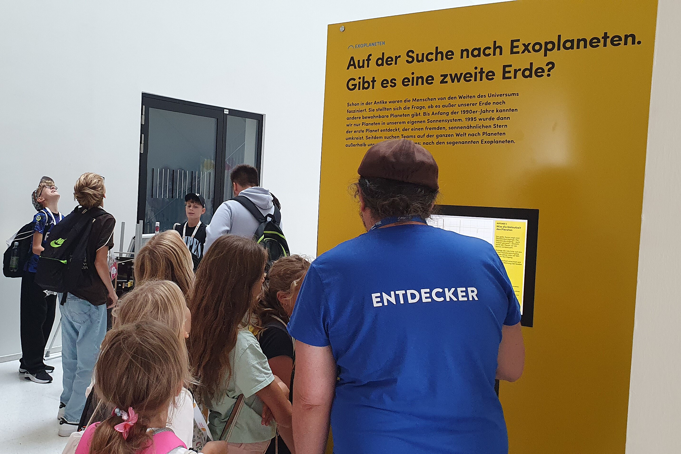 A group of children is standing in a brightly lit museum room in front of a yellow information panel about exoplanets. A staff member wearing a blue T-shirt with the word “EXPLORER” on it is explaining the exhibit to them. In the background, other visitors are standing next to an informational banner.