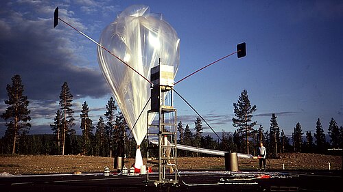 A large, transparent research balloon is being inflated at an outdoor launch site. In the foreground stands a tall metal frame equipped with measuring instruments and antenna masts jutting out at a wide angle, set against a backdrop of coniferous trees.