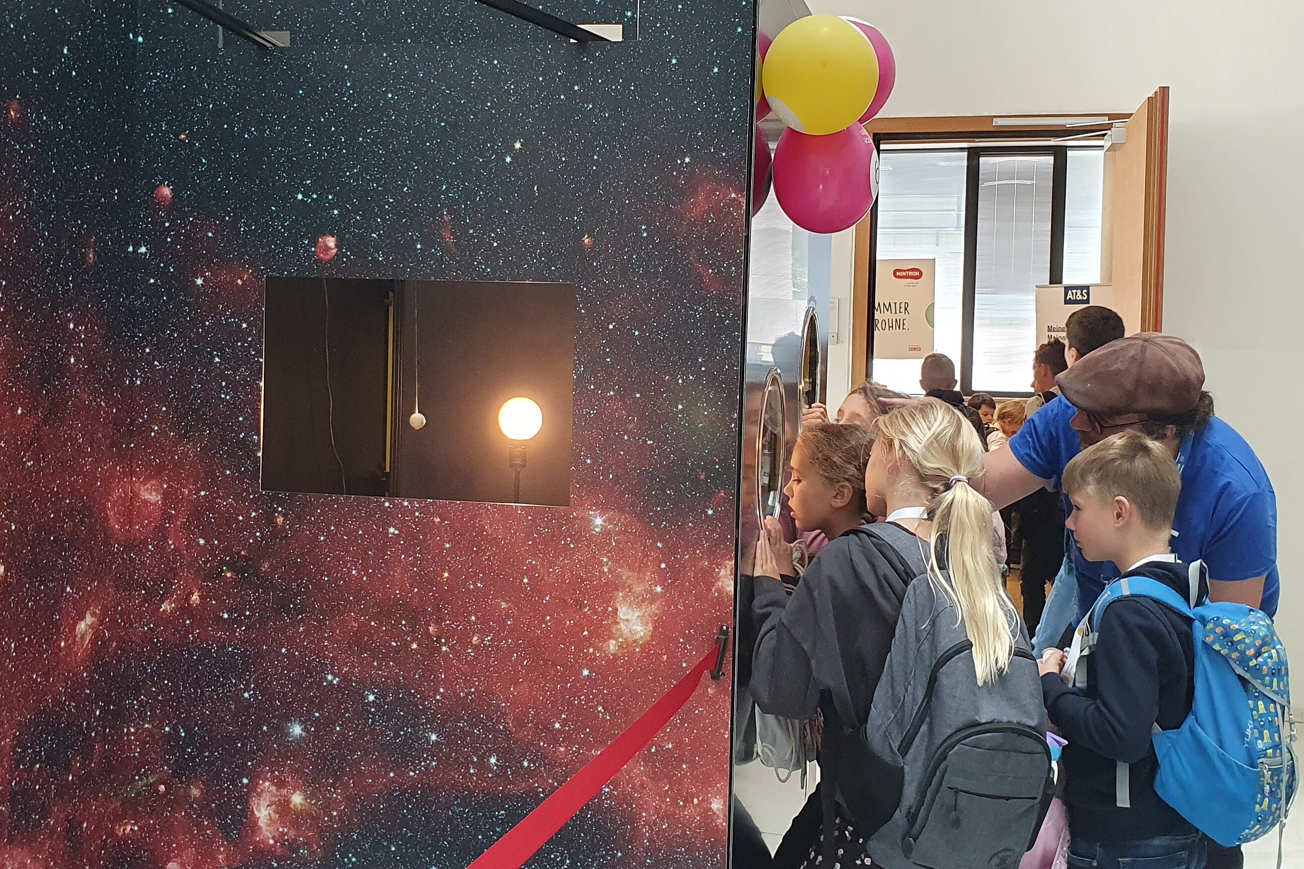 A group of children looking inside a box through a small window, while an adult explains to them what they see.