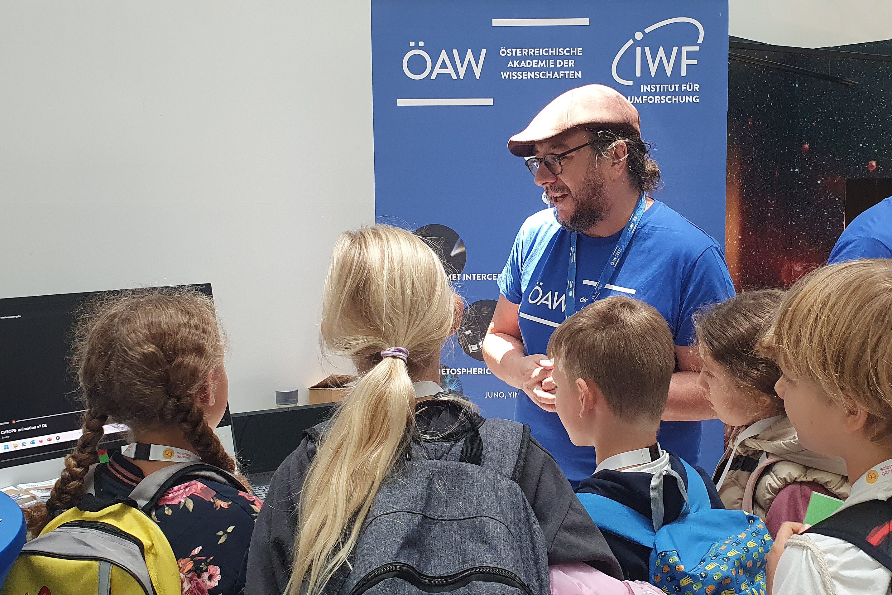 The image shows a group of kids looking at a man wearing a blue "ÖAW" T-Shirt, who is going to show them a video on a computer screen.