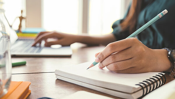 Eine Person sitzt am Schreibtisch und schreibt mit der einen Hand mit einem Stift in ein Notizbuch, mit der anderen Hand tippend am Laptop.