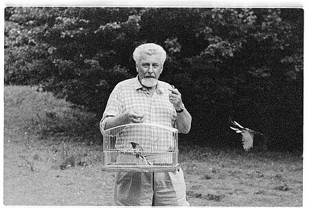 Konrad Lorenz steht im Freien und hält einen Vogelkäfig in den Händen, während ein Vogel aus dem geöffneten Käfig davonfliegt.