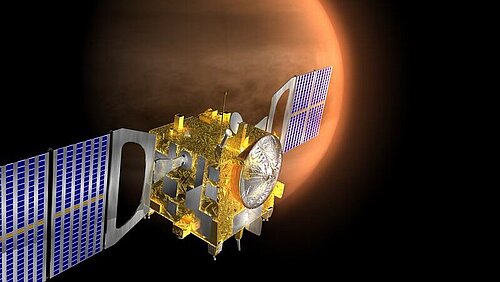 A cube-shaped, gleaming golden satellite with two broad, blue solar panel wings floats in the dark expanse of space. In the background, the crescent-shaped, densely clouded atmosphere of the planet Venus is visible.