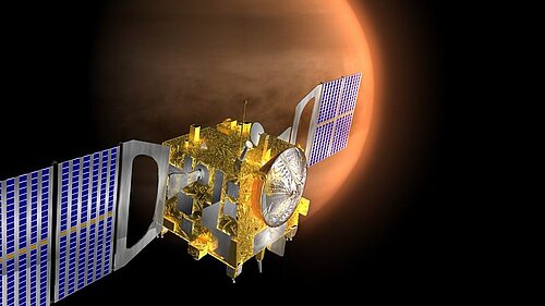 A cube-shaped, gleaming golden satellite with two broad, blue solar panel wings floats in the dark expanse of space. In the background, the crescent-shaped, densely clouded atmosphere of the planet Venus is visible.