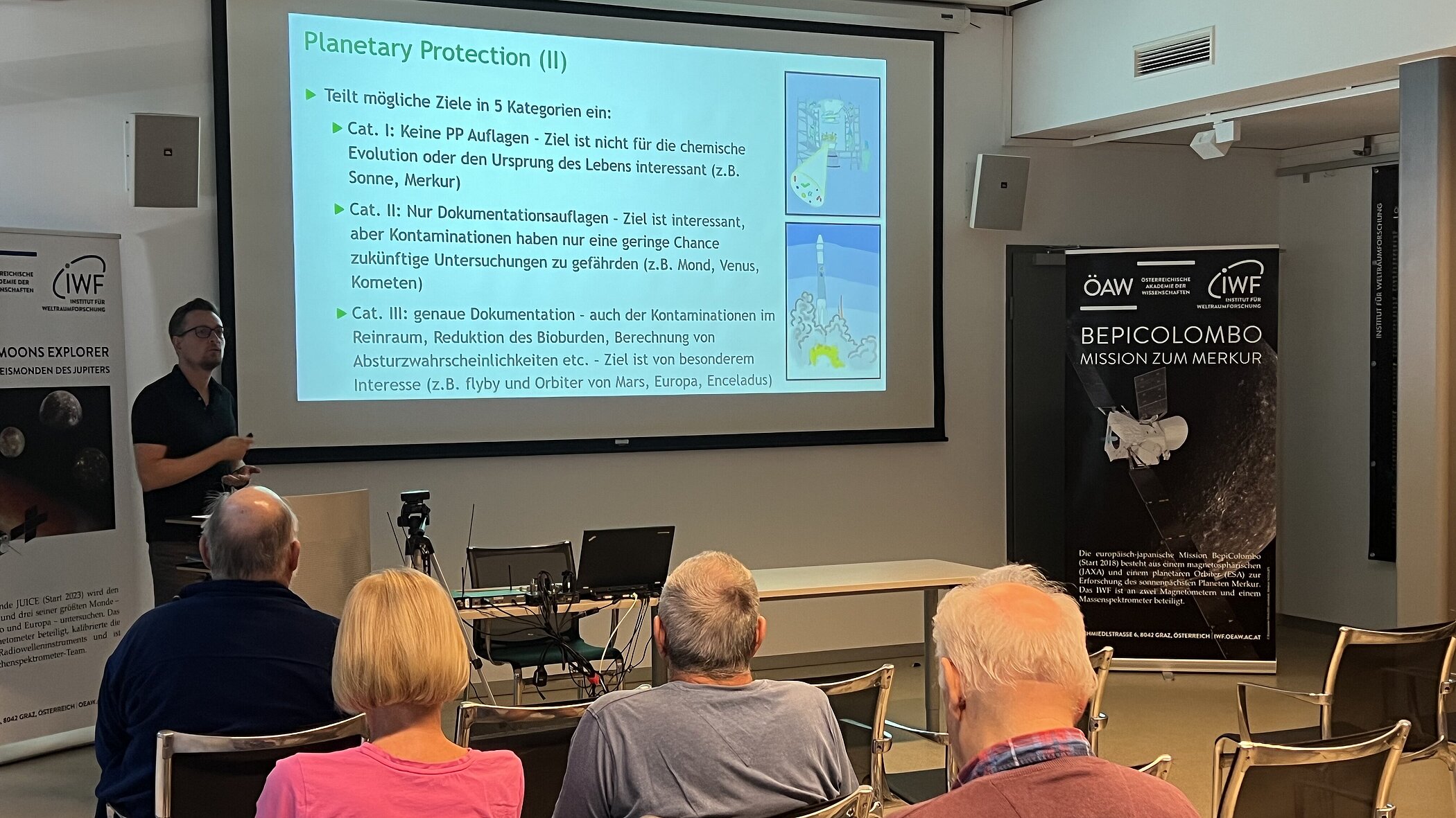 A speaker stands to the left in front of a large screen, giving a presentation on “Planetary Protection,” while the audience in the foreground is seen from behind, listening attentively. To the right of the screen stands a black promotional display for the “BepiColombo” space mission, featuring the logos of the Austrian Academy of Sciences (ÖAW) and the IMF.