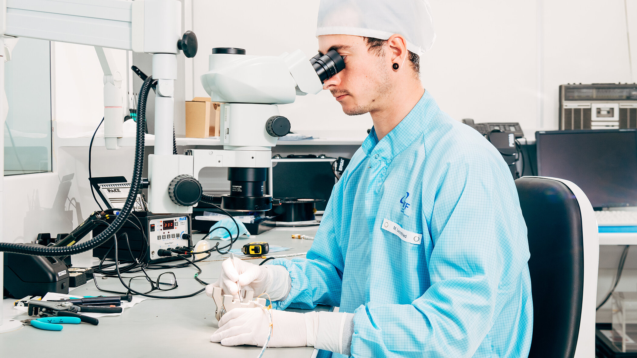 A researcher wearing a light blue lab coat and a white lab cap is intently looking through a large laboratory microscope. He is wearing white gloves and is working with tools on a small component on his clean workbench.