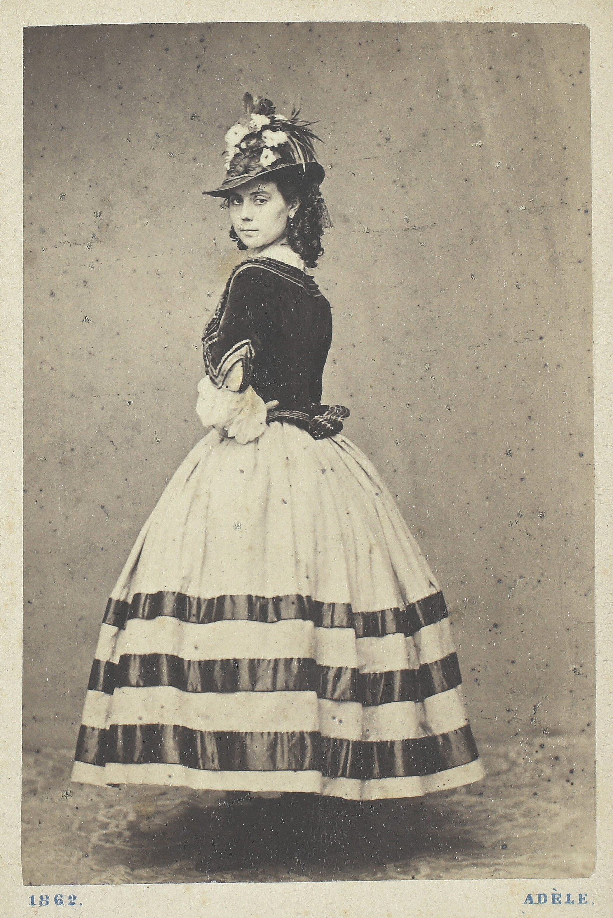 Ganzkörperportrait von Fräulein Schwöder. Sie ist halb von hinten abgebildet und schaut über ihre Schulter in die Kamera. Sie trägt einen Hut und ein Kleid mit breitem Rock und drei Streifen.