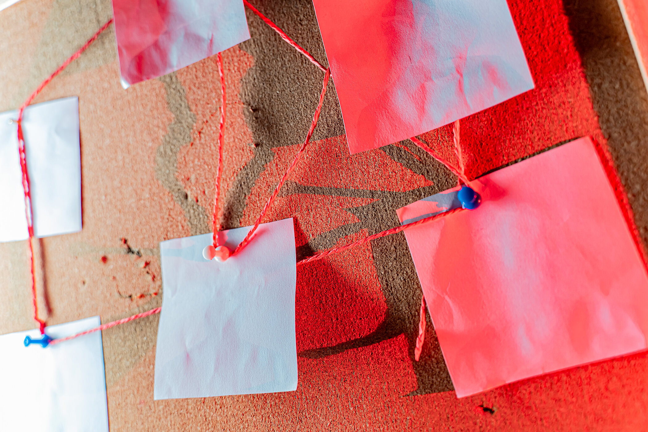 An evicence board made from cork, with little paper notes pinned to it, connected with red thread. The notes are empty and there is red lighting coming from the side.