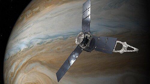 The Juno space probe is flying through space in front of the giant, striped planet Jupiter. The probe has a round body and three long, outstretched solar panels arranged like a three-bladed propeller. 