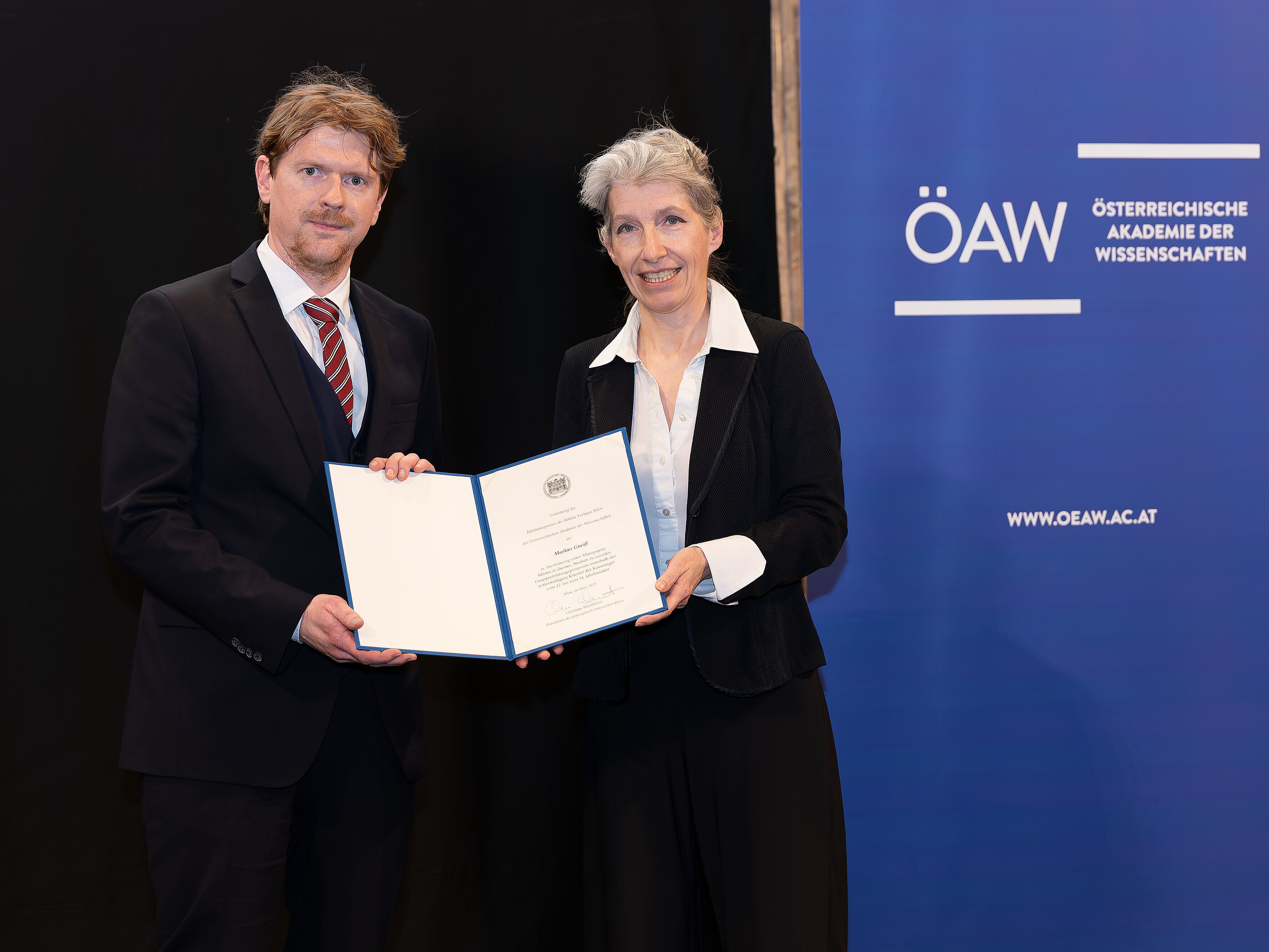 Preisträger Markus Gneiß und Christiane Wendehorst, Präsidentin der philosophisch-historischen Klasse bei der Übergabe der Urkunde; im Hintergrund das ÖAW-Logo