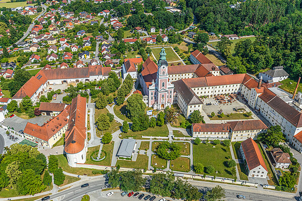 Das Kloster Aldersbach in Vogelperspektive. Es besteht aus mehreren Gebäudetrakten, einer Kirche und einem Park. Es gibt auch einen Innenhof und einen Parkplatz. Die Ortschaft ist hinter dem Kloster zu sehen und geht rechts in einen Wald über.