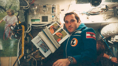 Austrian cosmonaut Franz Viehböck, wearing a blue jumpsuit and holding a flag, looks into the camera from inside a cramped space station filled with equipment and cables. In his hands, he holds an open manual with illustrations.
