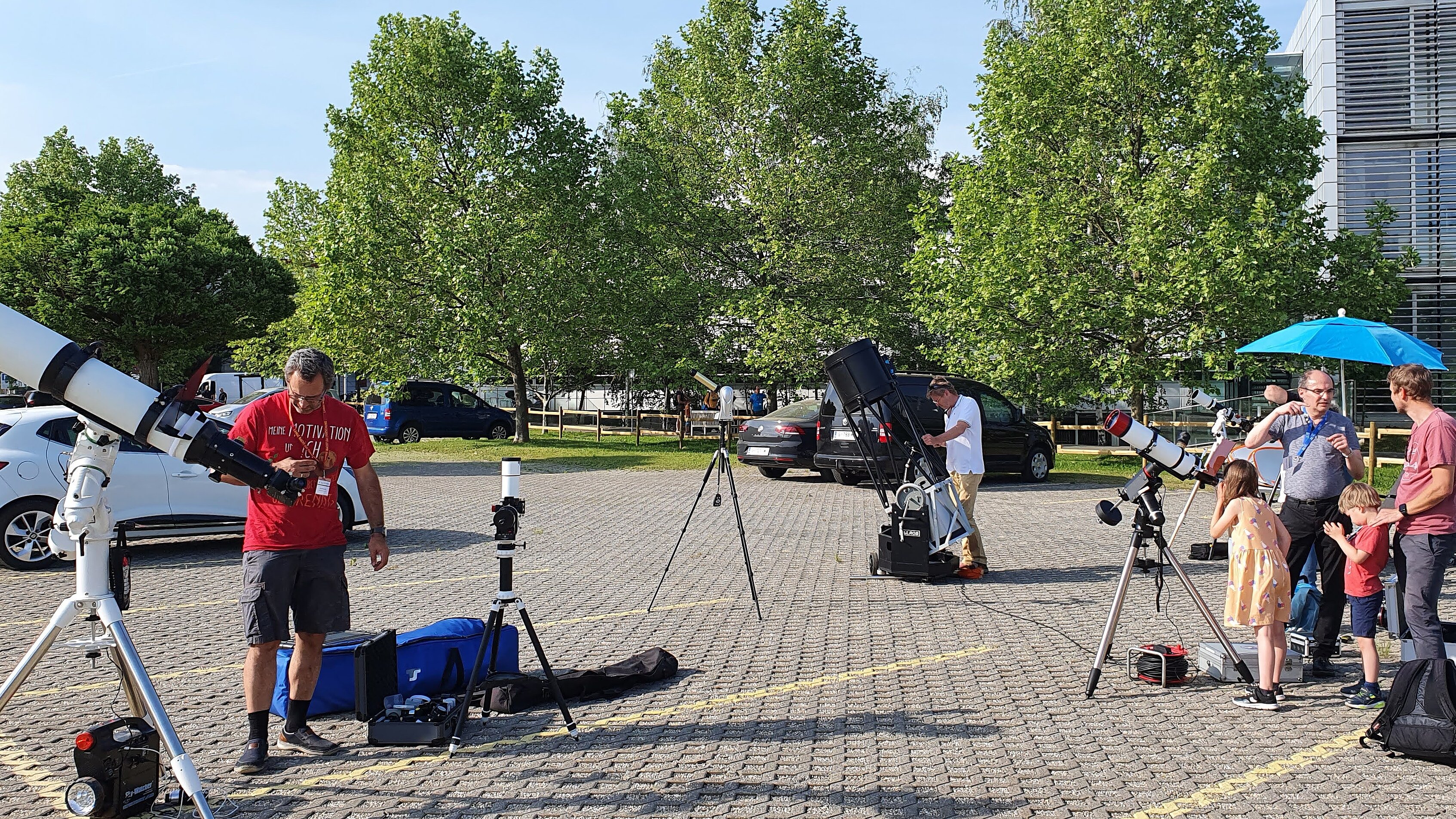An exterior shot taken on a sunny day shows several people and children in a paved parking lot setting up and adjusting various large telescopes for stargazing. In the background, green trees, parked cars, and a modern building with blinds are visible under a blue sky.