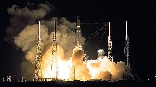The picture shows the Cassini/Huygens space probe being launched. A large cloud of smoke has formed around the probe, and four struts are attached to the rocket with steel cables.